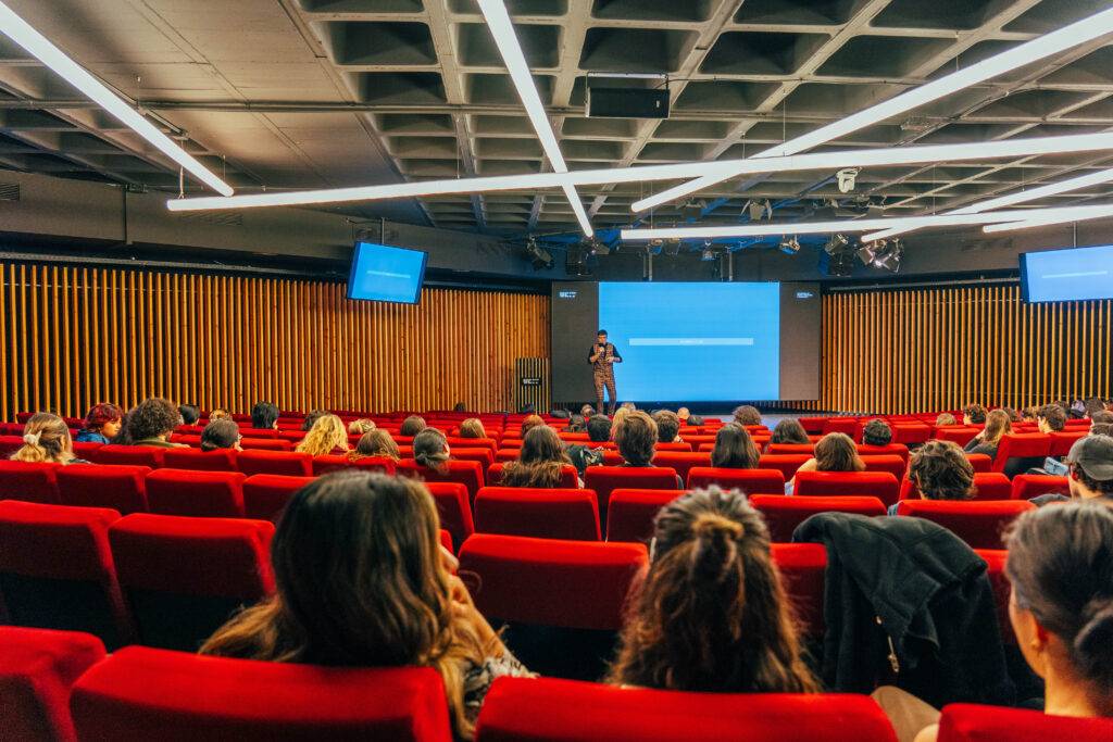 Una conferencia o presentación en un auditorio con asistentes sentados en filas rojas.
