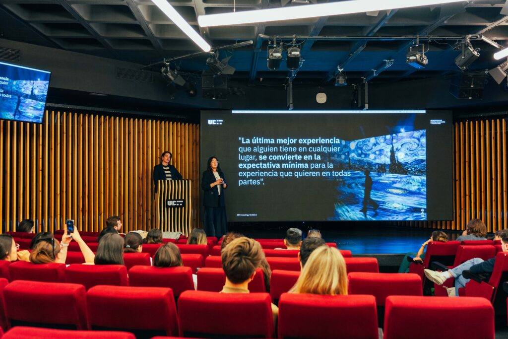 Una presentación en un auditorio con personas sentadas y dos oradores en el escenario.
