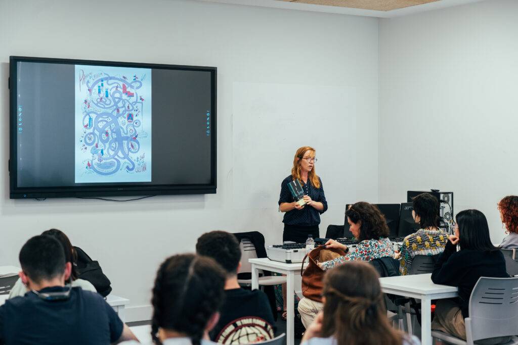 Una mujer presenta una ilustración en una pantalla mientras un grupo de estudiantes la observa atentamente.