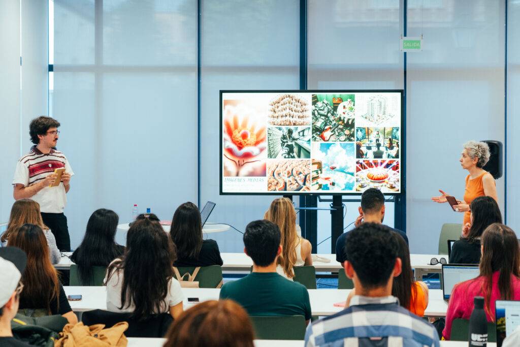 Una presentación en un aula con un expositor y una presentadora frente a una pantalla grande mostrando imágenes.