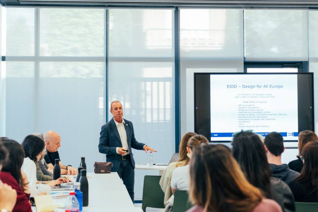 Un hombre presenta en una sala frente a un grupo de personas mientras se proyecta una presentación en la pantalla.