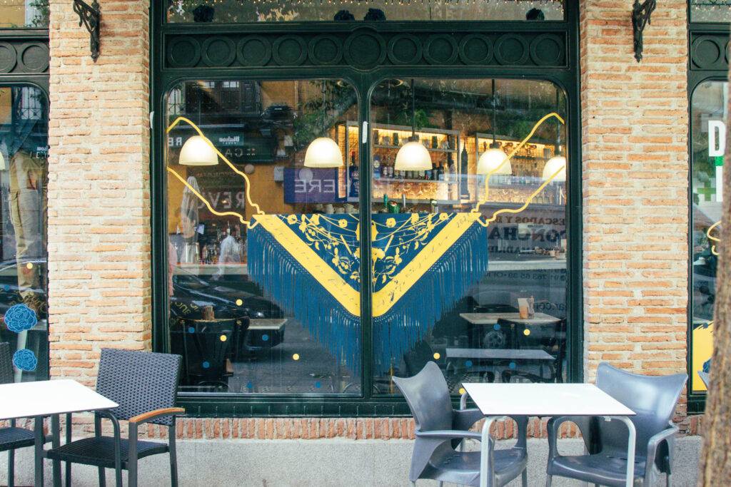La imagen muestra una ventana decorada con un diseño colorido de un mantón y mesas en el exterior.