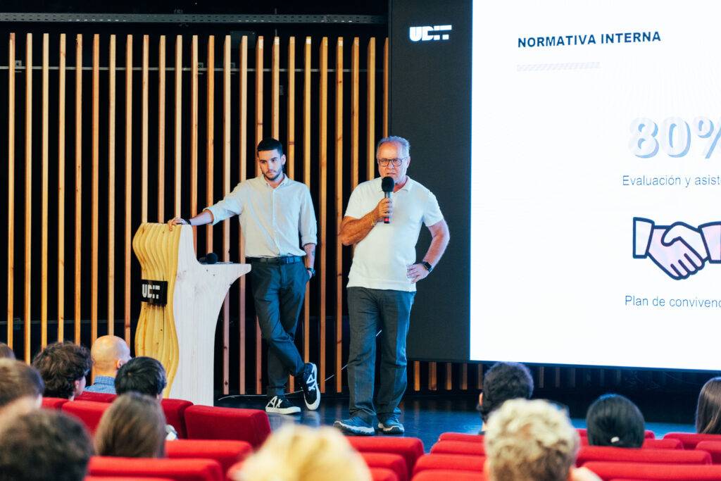 Una presentación en un auditorio donde dos hombres hablan frente a una pantalla con información.