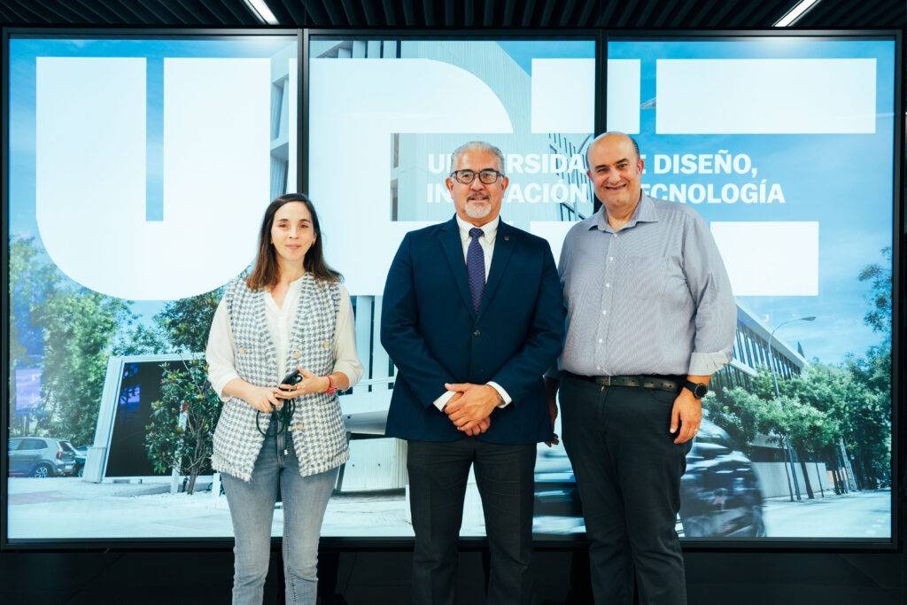 Tres personas posando frente a una pantalla con el logotipo de una universidad.