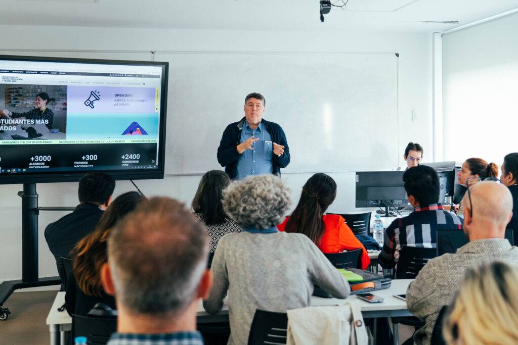 Una presentación en un aula con un orador frente a un grupo de asistentes que observan una pantalla grande.
