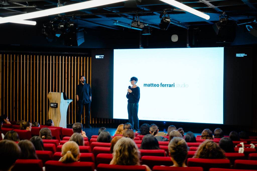 Un presentador habla ante un público en un auditorio moderno con asientos rojos.