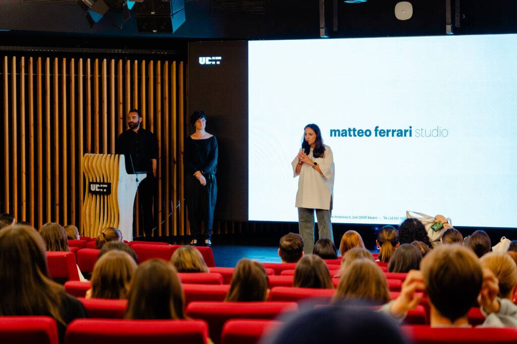 Una presentación en un auditorio con tres personas en el escenario y un público atento.