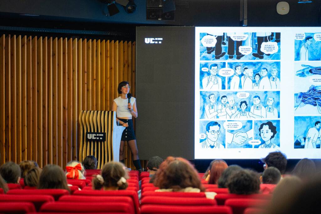 Una presentadora está en un escenario frente a una audiencia, mostrando una presentación de cómic en una pantalla grande.
