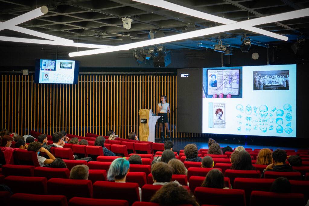 Una presentadora expone en un auditorio mientras el público escucha atentamente.