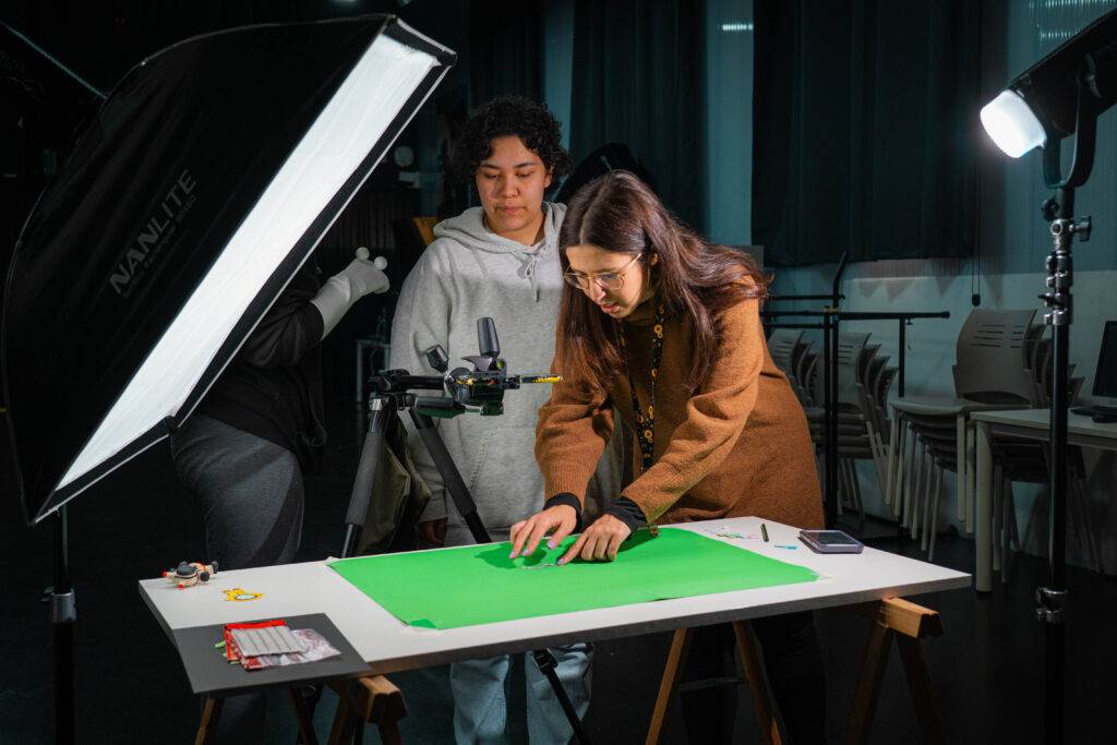 Dos personas están trabajando en un entorno de estudio con fondo verde y luces de fotografía.