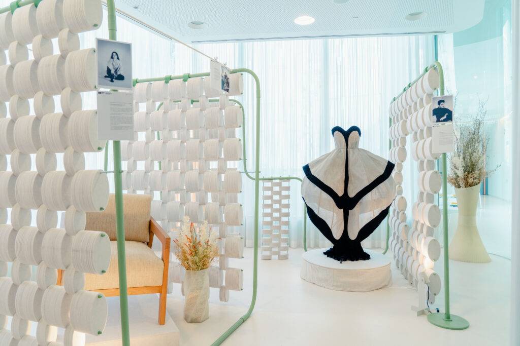 La imagen muestra una exhibición artística con una prenda destacada en el centro, rodeada de elementos decorativos y un ambiente moderno.