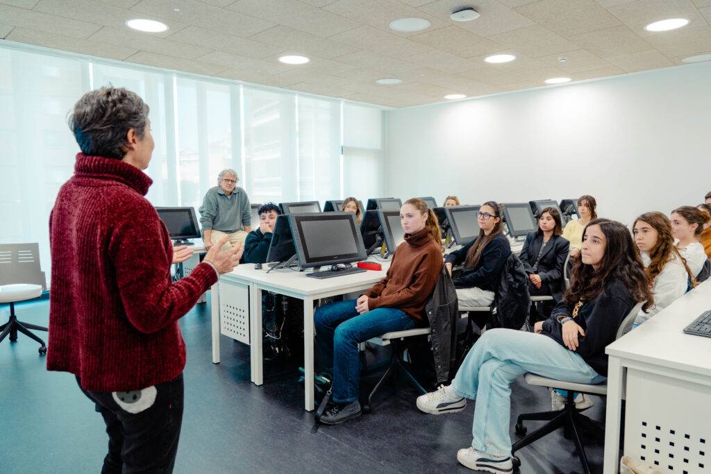 Un grupo de estudiantes escucha atentamente a un docente en un aula con computadoras.