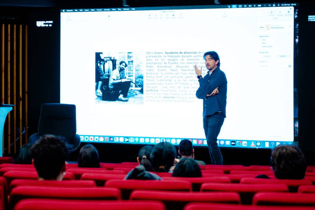 Un ponente presenta información en una pantalla grande mientras un público atento observa desde butacas rojas.