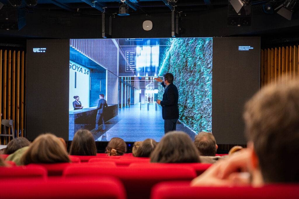 Una presentación en un auditorio moderno con una pantalla grande y vegetación decorativa.