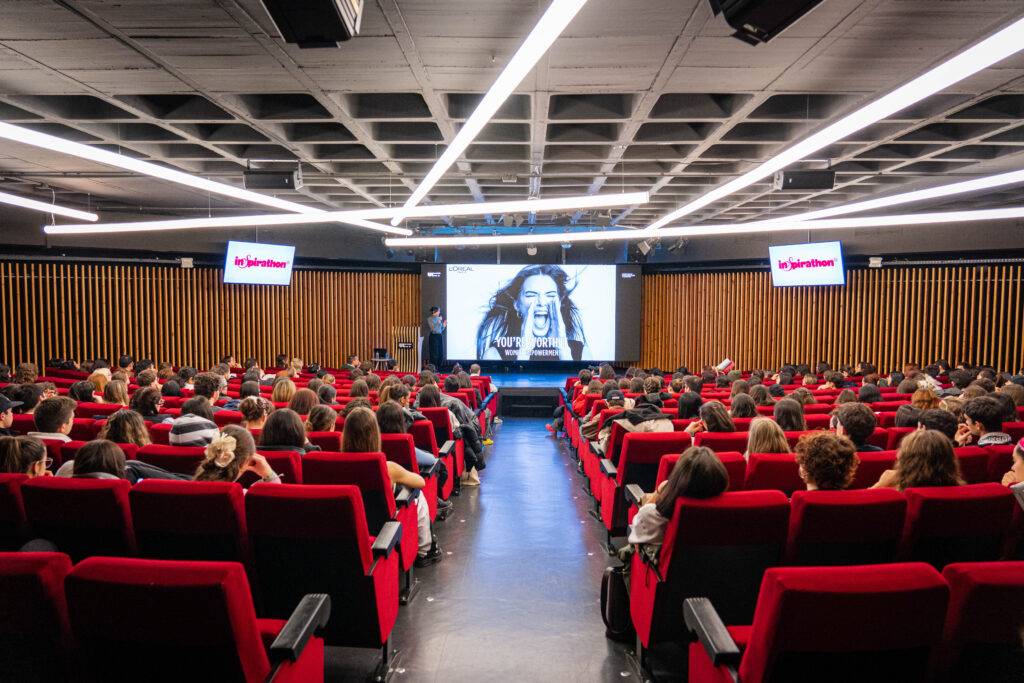 Una sala llena de personas sentadas frente a una pantalla grande durante una presentación.