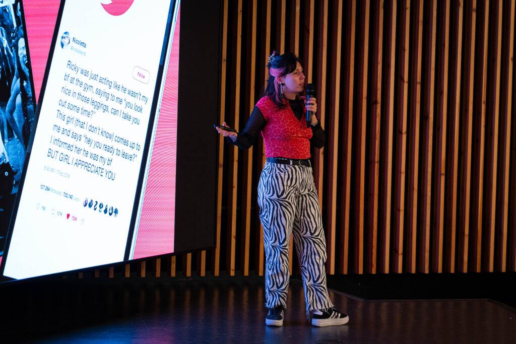 Una persona presenta en un escenario con una proyección de un tweet detrás.