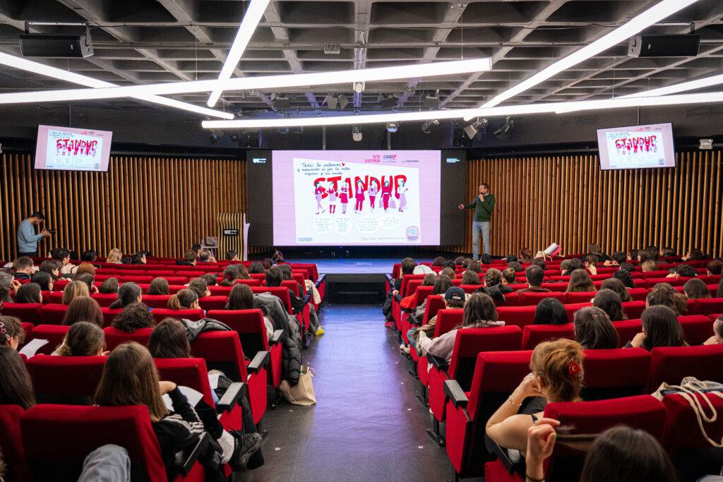 Una sala de conferencias llena de personas sentadas en filas rojas, mientras un ponente presenta en una pantalla grande.