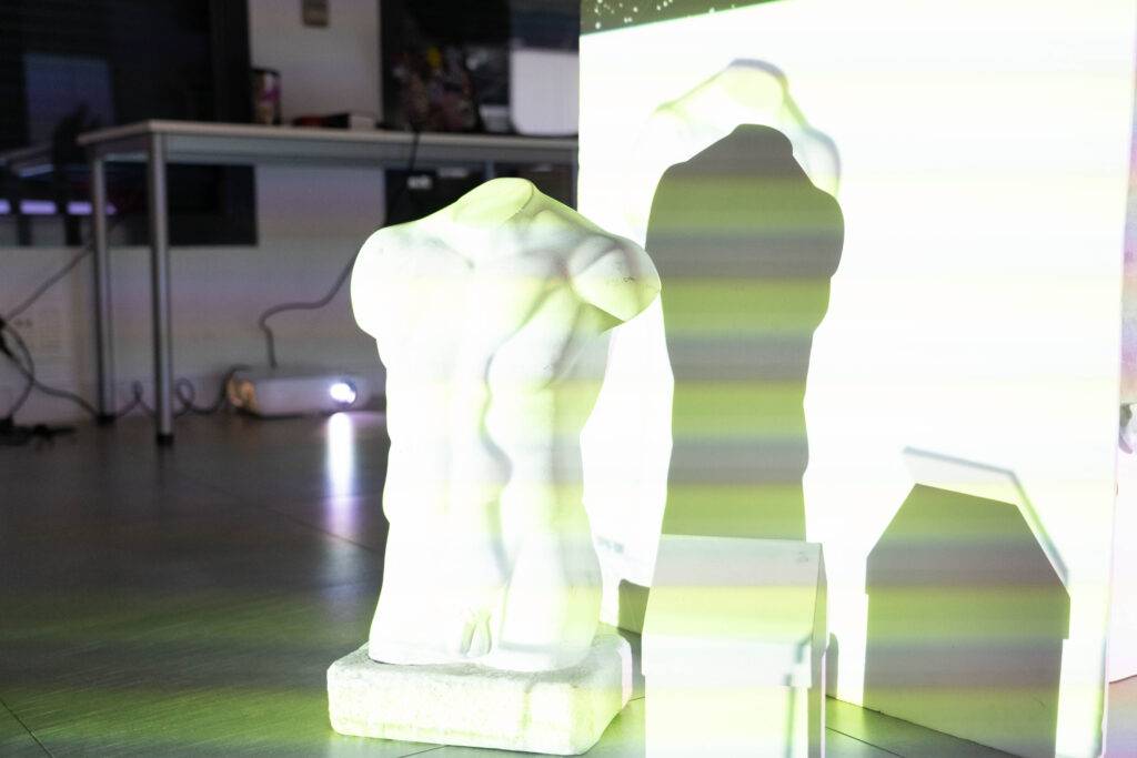 Una escultura de un torso humano iluminada con luces que crean sombras en la pared.