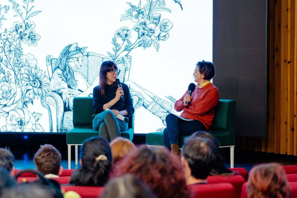 Dos mujeres conversan en un evento, con un fondo artístico de flores y una figura sentada.