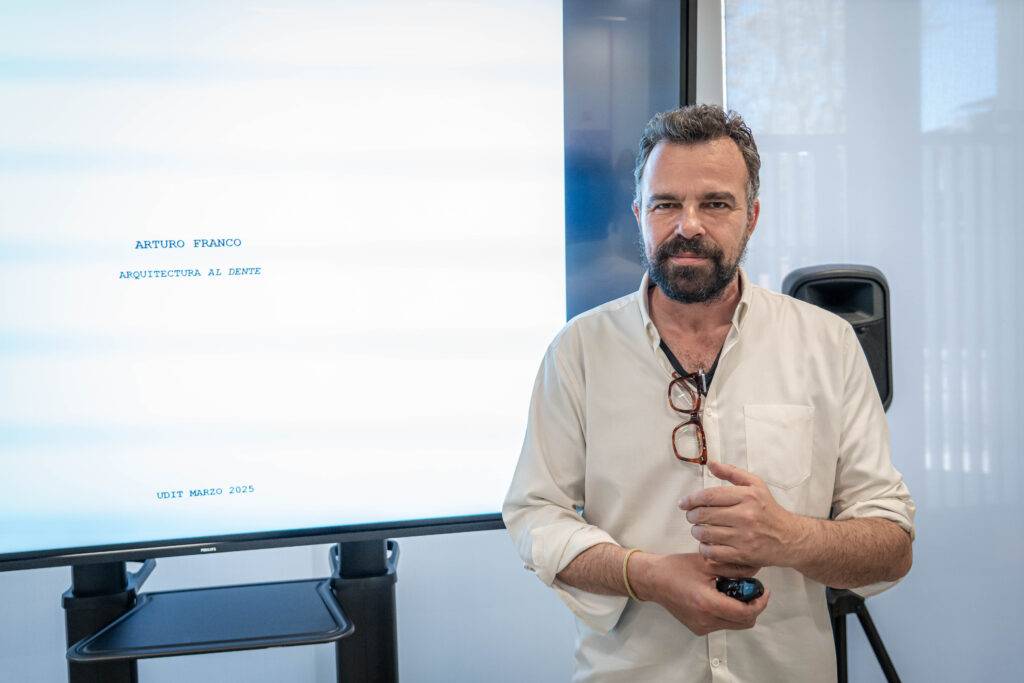 Un hombre de barba y camisa clara está presentando frente a una pantalla.