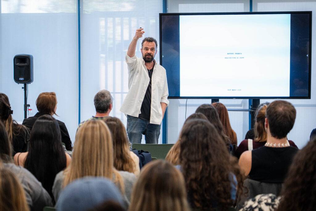 Un hombre en una presentación sostiene un objeto mientras se dirige a una audiencia sentada frente a una pantalla.