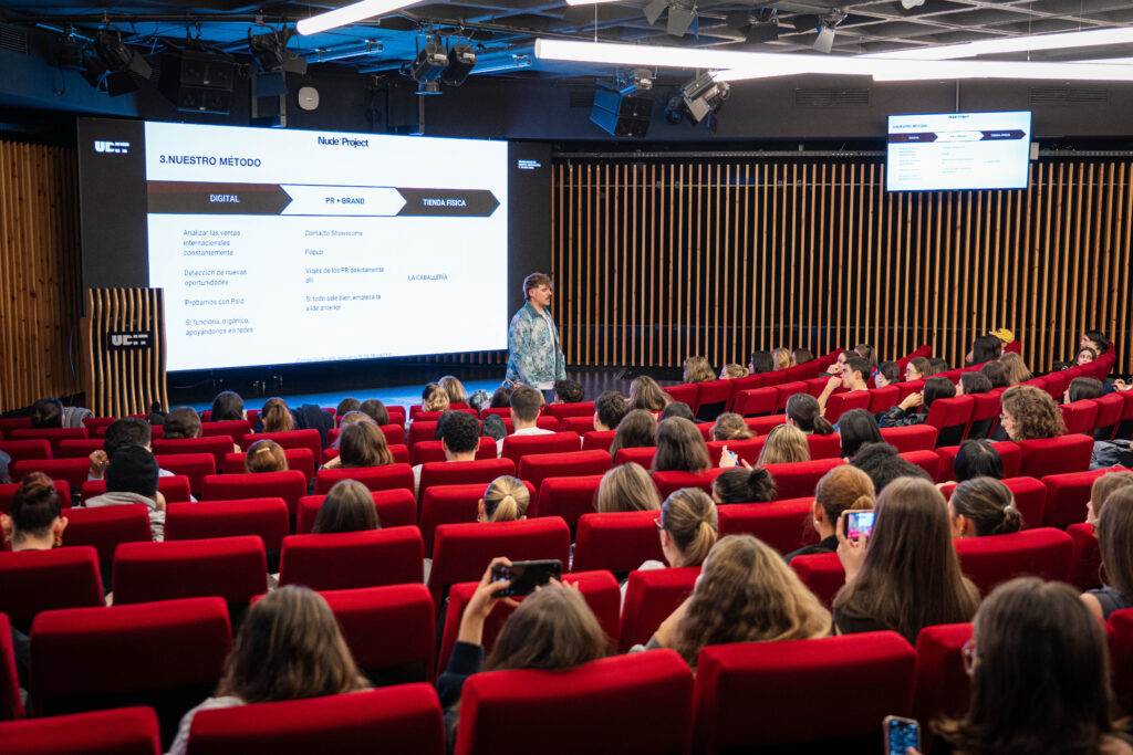 Una charla en un auditorio con un orador frente a una audiencia sentada en asientos rojos.