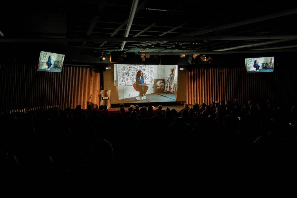 Una presentación en un auditorio con una audiencia atenta y una pantalla mostrando una persona sentada.