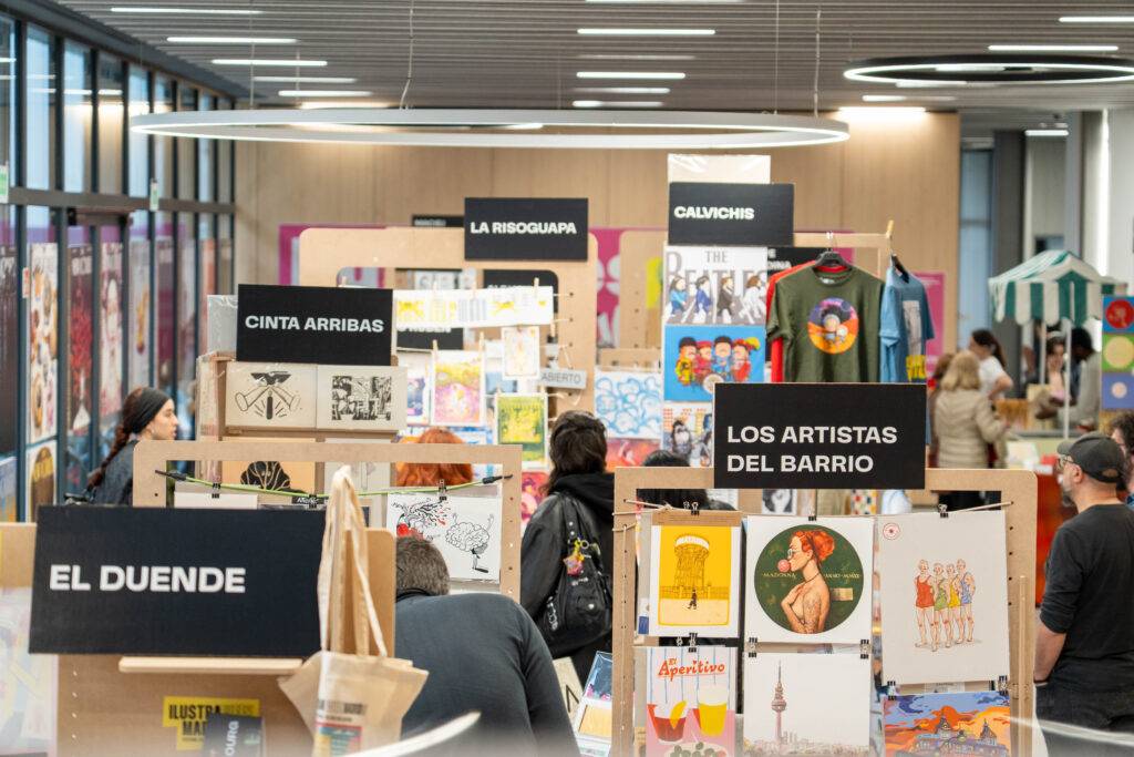 Una feria de arte en un espacio interior donde se exhiben obras y carteles de diferentes artistas.