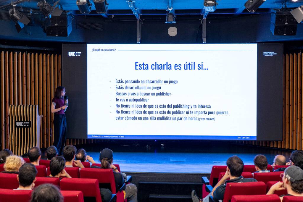 La imagen muestra a una presentadora hablando sobre el desarrollo de videojuegos frente a un público atento en un auditorio.