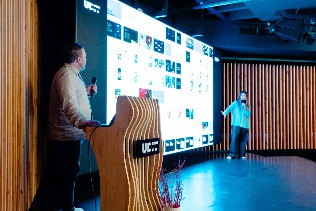 Una presentación en un escenario con una pantalla grande y dos oradores.