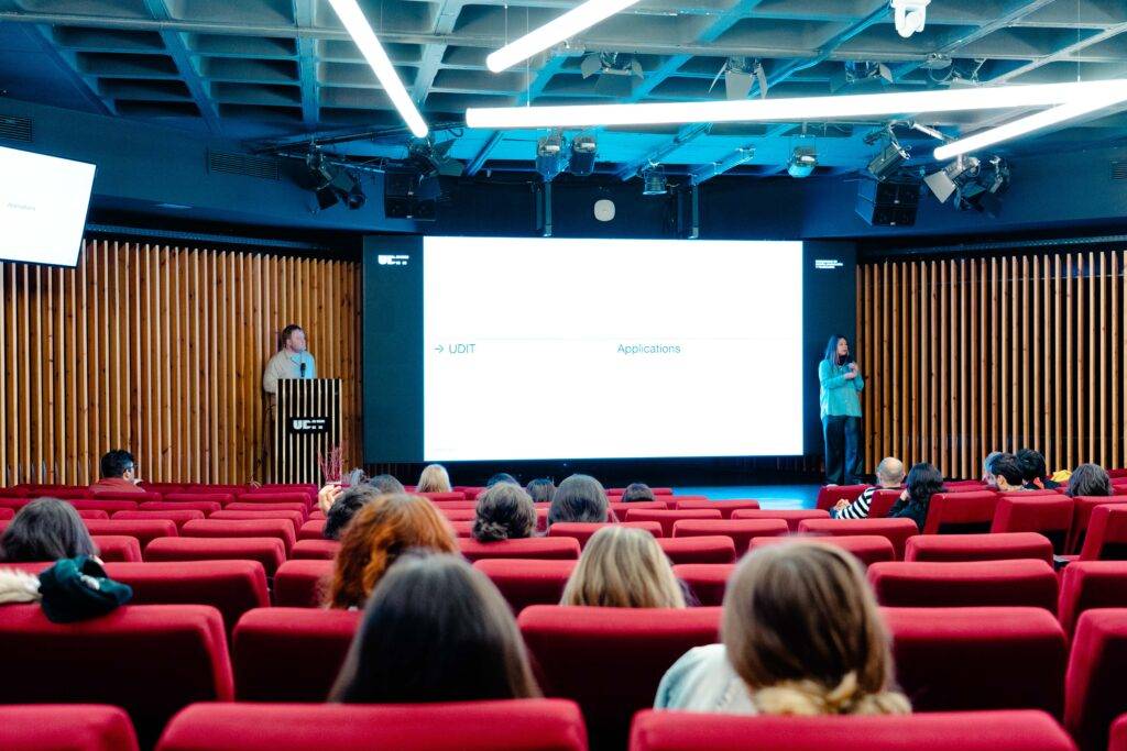 La imagen muestra a dos presentadores hablando frente a una audiencia en un espacio de conferencias moderno con asientos rojos.