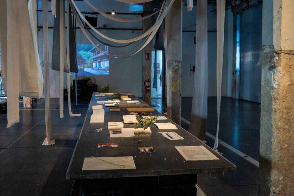 Un espacio expositivo con una mesa central llena de papeles y una decoración de cintas colgantes.