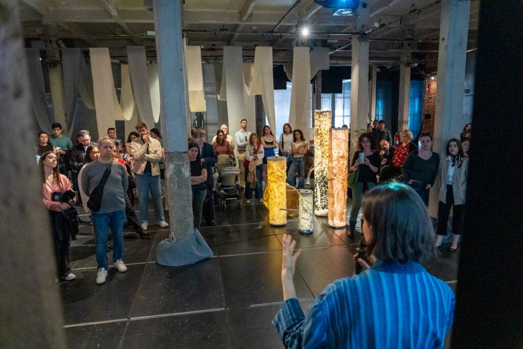 Un grupo de personas observa a una oradora en un evento en un espacio artístico.