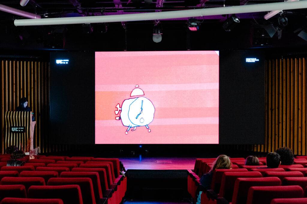 Una presentación audiovisual con un reloj animado en una pantalla y un público sentado frente a ella.