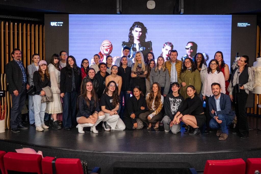 Un grupo de personas posando juntas en un escenario con una pantalla grande detrás que presenta imágenes de moda.