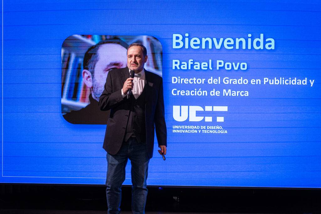 Un hombre de pie en el escenario dando la bienvenida a un evento académico.