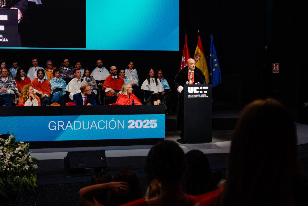 Una ceremonia de graduación con un orador en un podio y un público con togas y birretes.