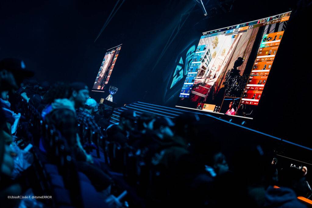 Una multitud observa una competición de videojuegos en un escenario iluminado con pantallas grandes mostrando la jugabilidad.