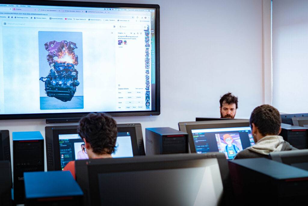 Una sala de clases con estudiantes trabajando en computadoras y una proyección en la pantalla frontal.