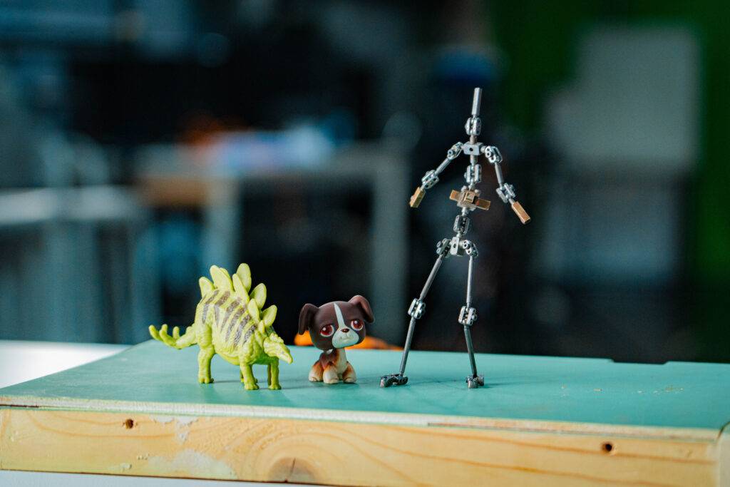 La imagen muestra un dinosaurio de juguete, un perro de peluche y una figura de modelo artístico sobre una superficie verde.