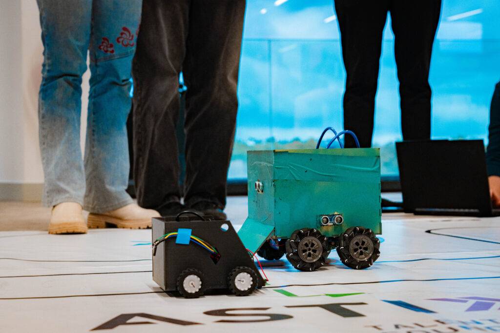 Dos robots pequeños están en una superficie con el logotipo de una empresa mientras varias personas están de pie alrededor.