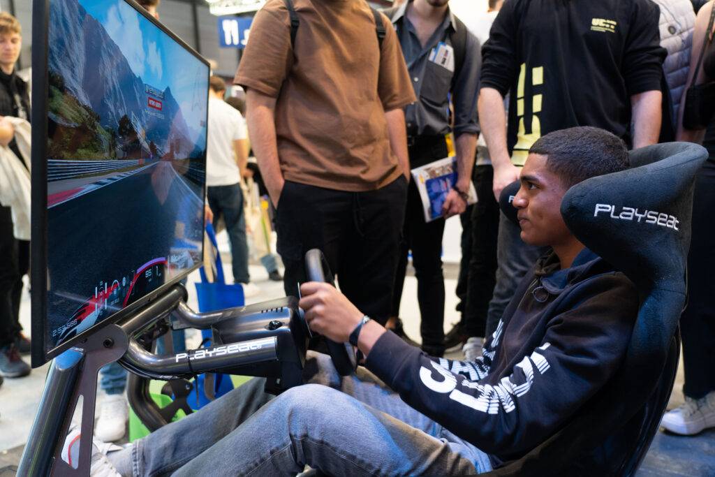 Un joven sentado en un simulador de carreras, concentrado en el juego mientras otros observan.