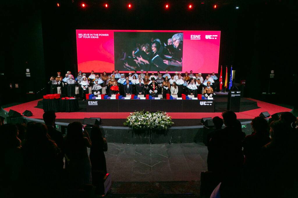 Ceremonia de graduación con estudiantes y facultad en un escenario iluminado.