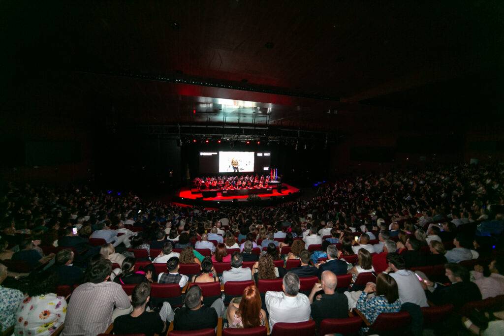 Una multitud de personas en un auditorio oscuro observando una presentación en el escenario.