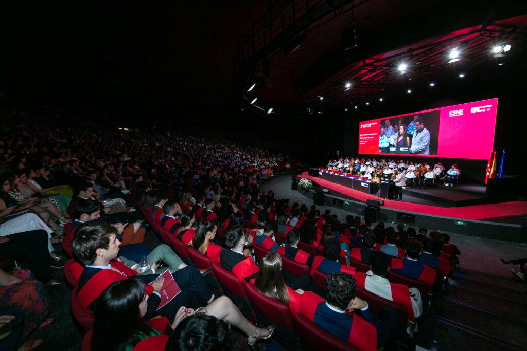 Una multitud de personas observa un evento en un auditorio con un panel en el escenario.