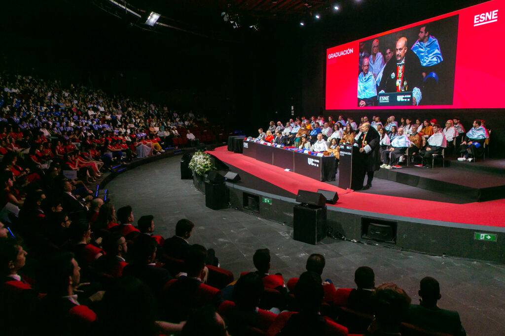 Ceremonia de graduación con un gran auditorio lleno de estudiantes y un presentador en el escenario.