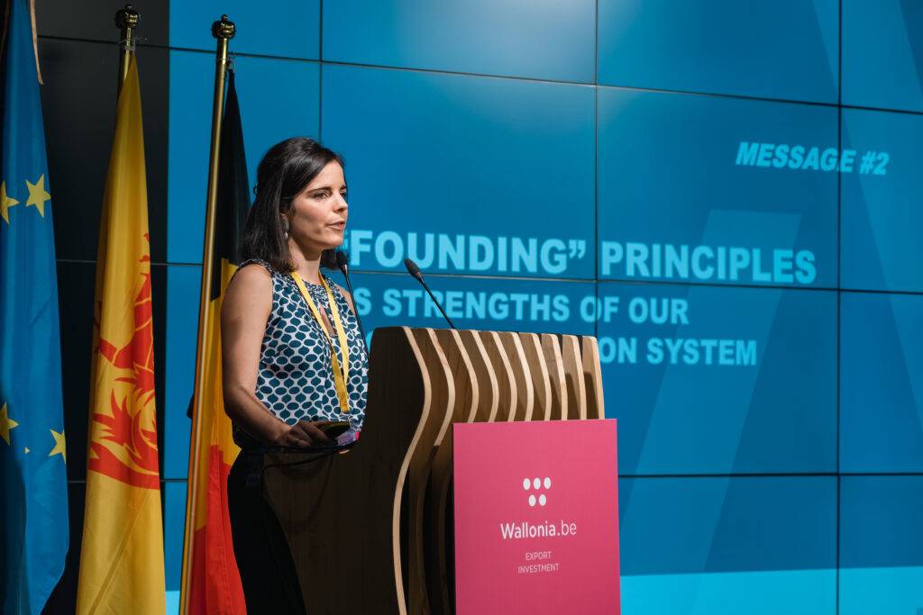 Una mujer presenta un discurso frente a un panel con banderas y una pantalla que muestra información sobre principios fundacionales.