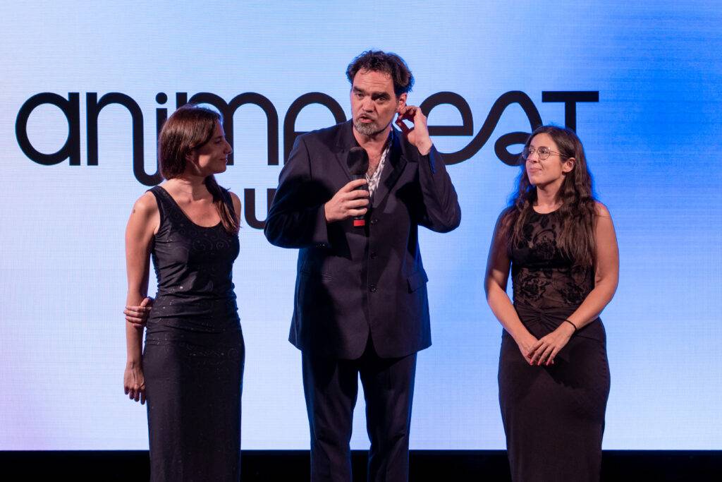 Tres personas en un evento, dos mujeres a un lado y un hombre en el centro con un micrófono.