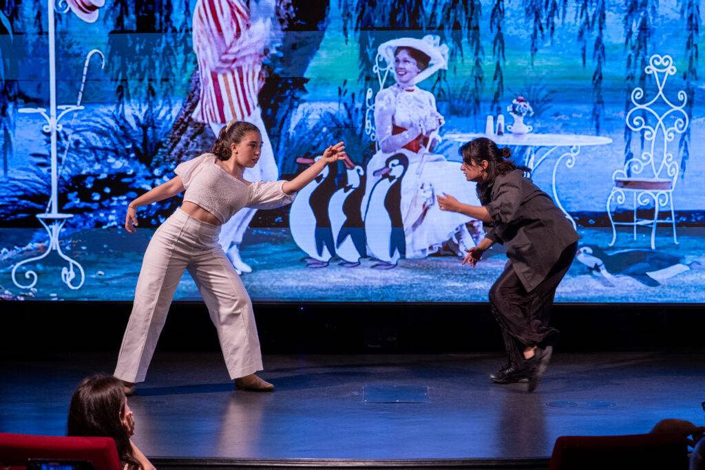 Dos bailarines se presentan en un escenario frente a una proyección colorida de un fondo con pingüinos.