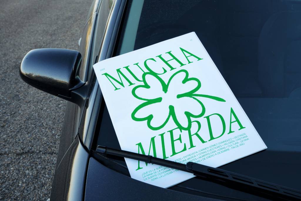 Una hoja con el texto 'MUCHA MIERDA' y un diseño de trébol verde está apoyada sobre el capó de un coche.